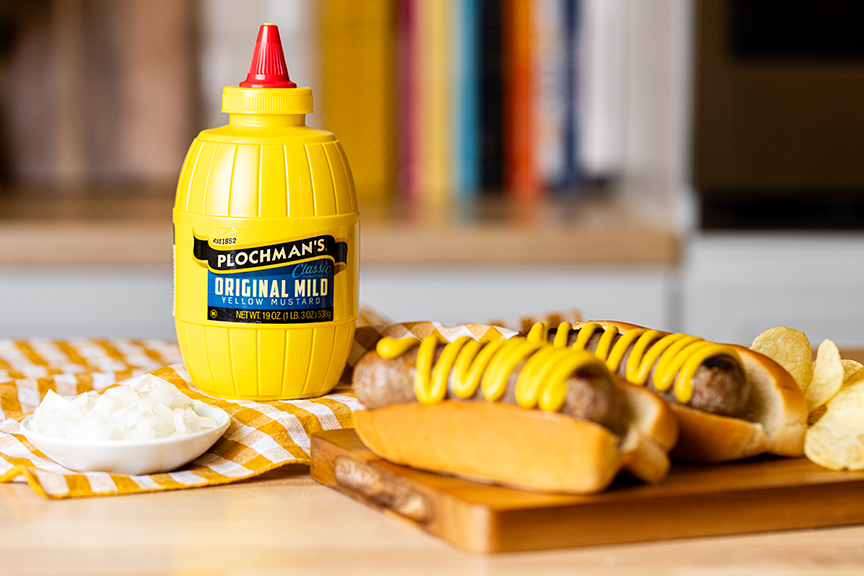 Yellow mustard bottle with red cap on a wooden cutting board with hot dogs and chips.